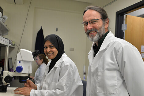 Two scientists in white coats smile by a microscope in a research lab